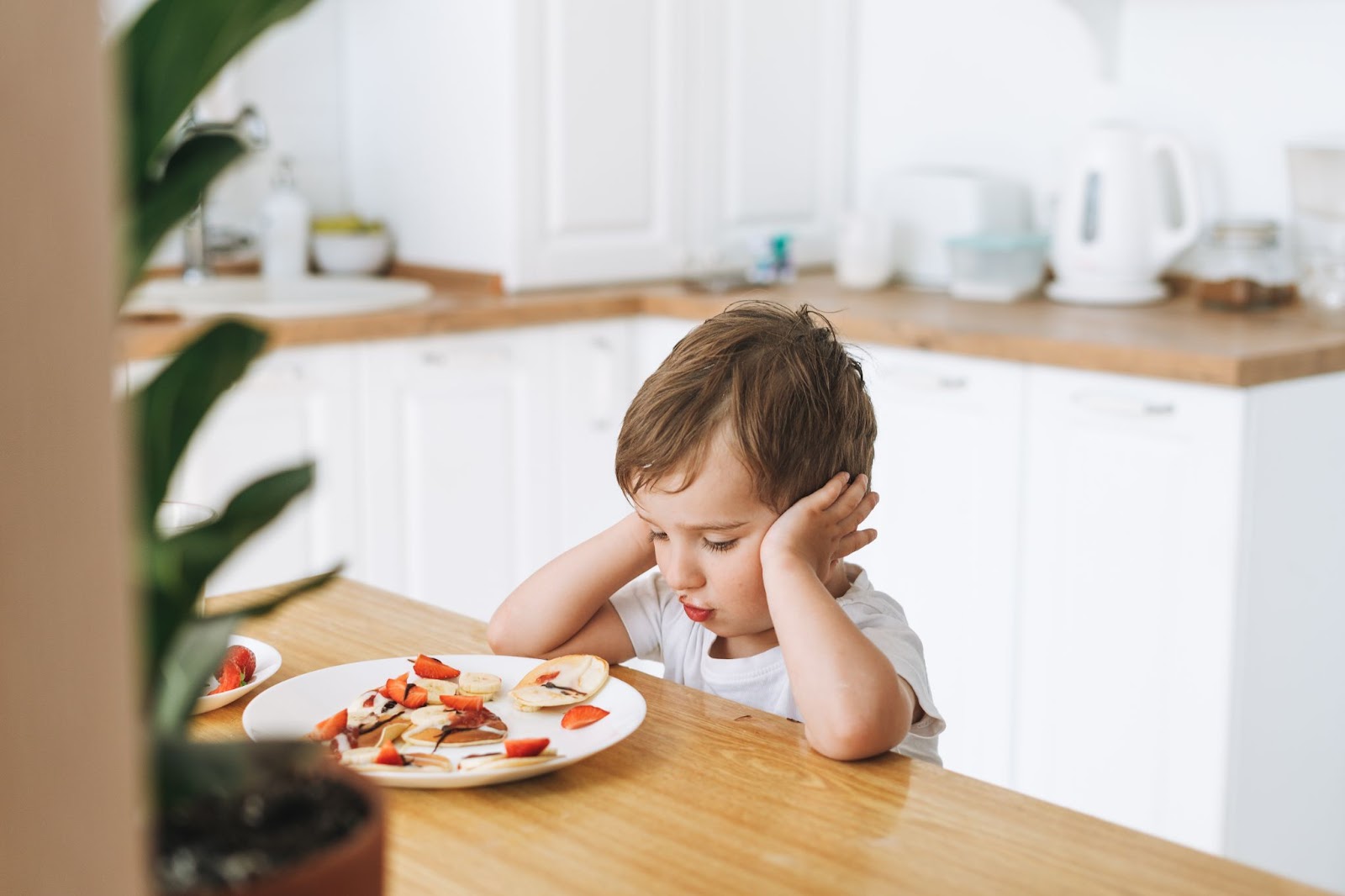 Bambino carino seduto con puncake con bacche in cucina luminosa a casa. Il bambino non vuole mangiare